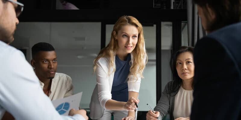 Confident skilled businesswoman explain marketing data paper report financial results analysis to multi-ethnic colleagues, company staff gathered at meeting brainstorming thinking strategizing concept